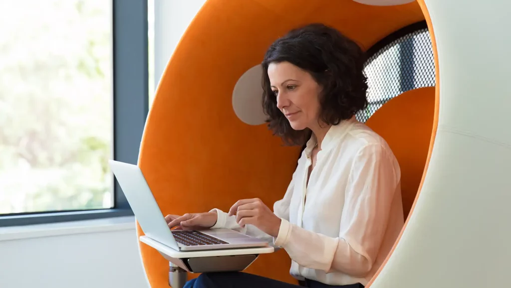 Mulher trabalhando em um computador laptop para recrutamento dentro de uma moderna poltrona circular laranja, refletindo o ambiente de trabalho contemporâneo e produtivo.