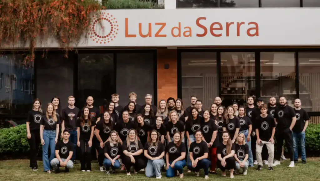Grupo de funcionários da Luz da Serra posando em frente à fachada da empresa, todos usando camisetas pretas com logotipo. Ambiente empresarial ao ar livre.