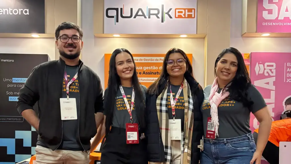 Grupo de quatro pessoas sorridentes participando de evento sobre gestão de pessoas no estande da Quark RH no evento CONARH 2025. empresa de recursos humanos, com destaque para banners e ambiente de feira.
