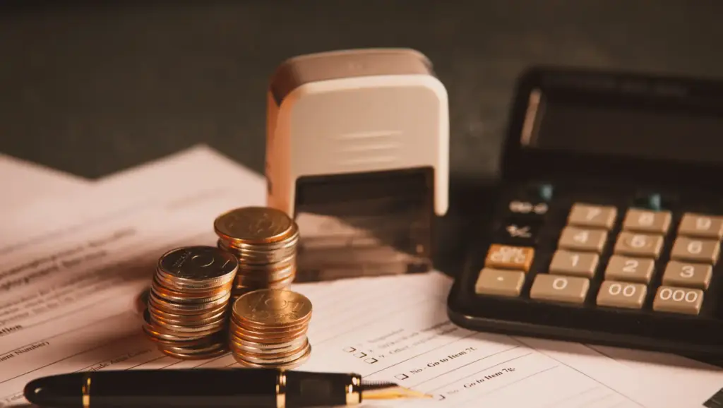 Imagem de moedas empilhadas, calculadora e documentos financeiros sobre uma mesa, representando previdência privada, gestão financeira, economia ou negócios.