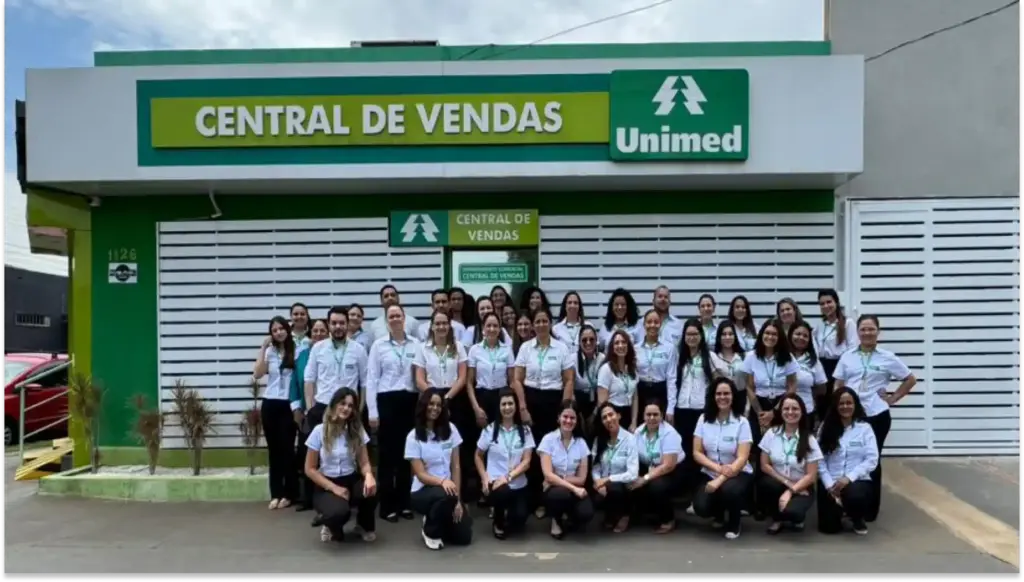 Grupo de profissionais sorridentes em ambiente corporativo, vestindo uniformes com logotipo e identidades diversas, promovendo colaboração e trabalho em equipe Unimed Três Lagoas