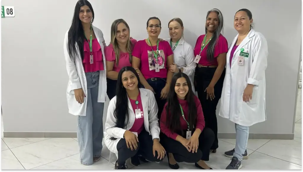 Grupo de profissionais de saúde em uma visita hospitalar, sorrindo para a câmera, com uniforme rosa e branco, demonstrando cooperação e cuidado.