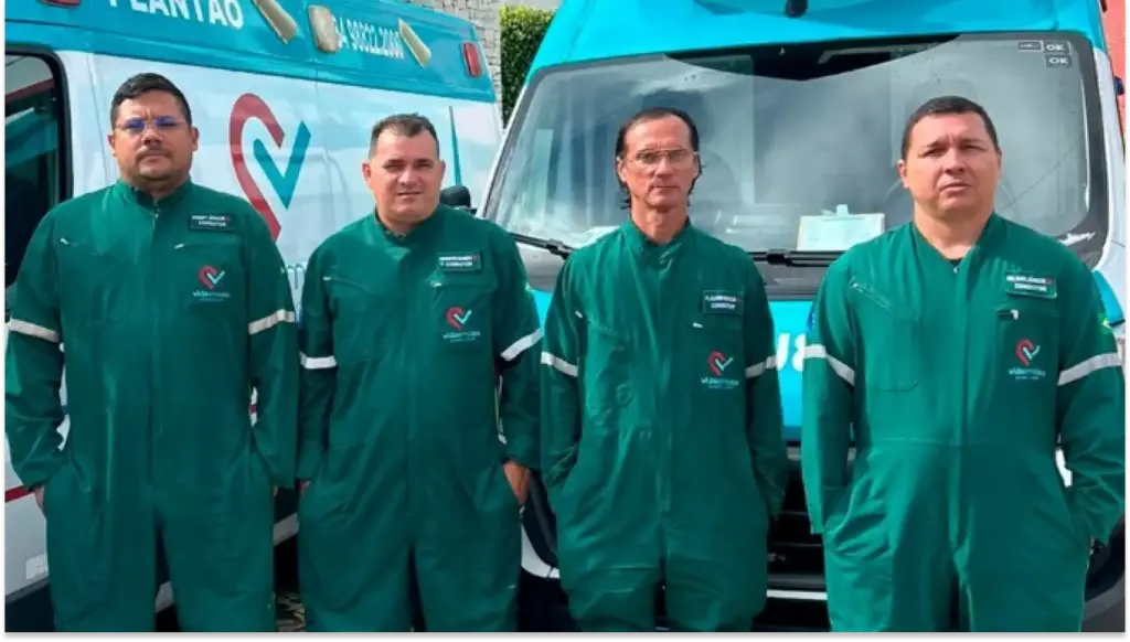 Equipe de profissionais de saúde posando ao lado de uma ambulância azul, usando jalecos verdes, representando serviço de emergência médica e saúde preventiva.