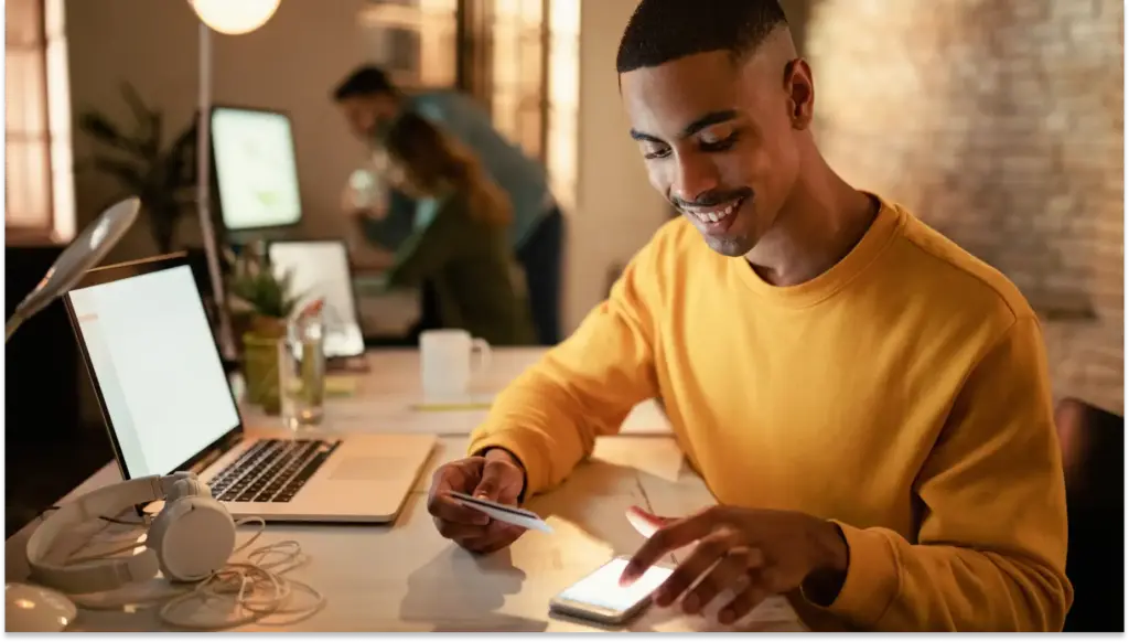 Homem sorridente usando smartphone em ambiente de coworking, com computadores, notebooks e pessoas ao fundo, criando um ambiente de trabalho colaborativo e recebendo a primeira parcela do 13° salário.