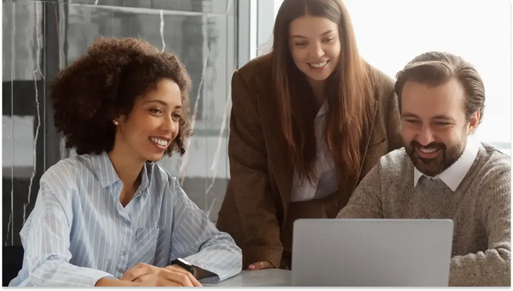 Três jovens profissionais de diferentes etnias sorrindo durante uma reunião de trabalho, usando roupas de escritório, em um ambiente moderno com janelas grandes ao fundo, focados em um laptop contendo o Relatório de Transparência Salarial.