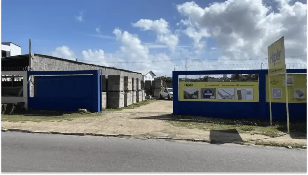 Foto de uma loja de materiais de construção chamada Hiper da construção, situada na rua com estacionamento ao lado e uma área de areia na frente, sob céu com nuvens.
