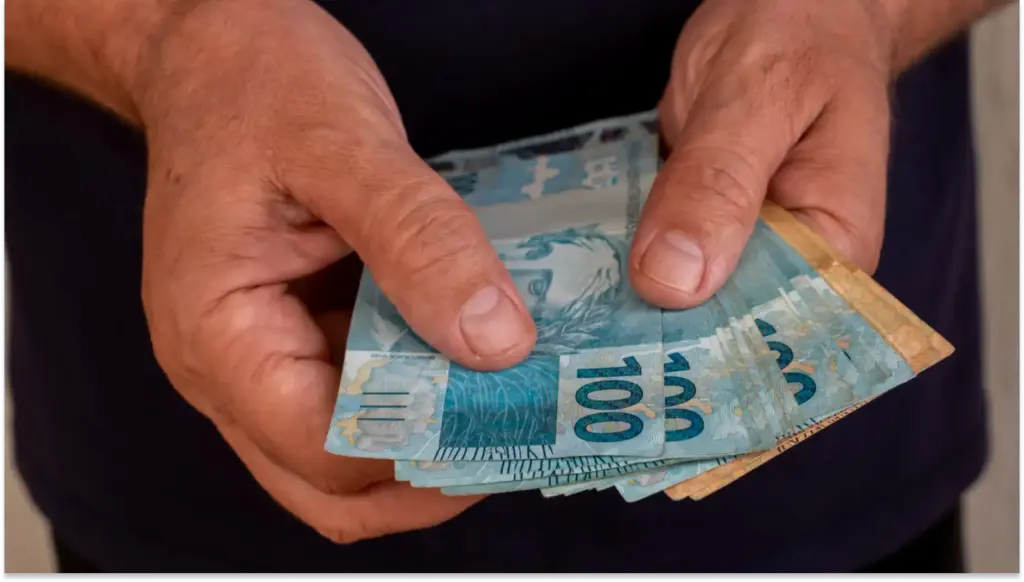 Mãos segurando várias notas de cem reais, destaque para a textura das cédulas e os detalhes amarelados na borda em seu saque-aniversário.