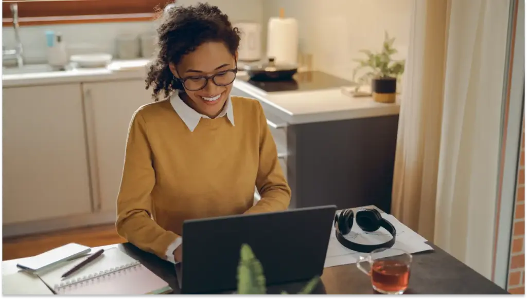 Mulher sorridente usando laptop em mesa de trabalho em casa, com notebooks, fone de ouvido e chá, ambiente acolhedor e bem iluminado fazendo trabalho híbrido 2.0.
