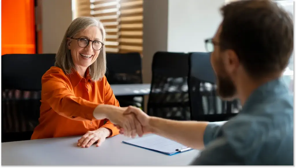 Mulher idosa sorridente dando um aperto de mão a um homem durante uma entrevista de emprego, ambos sentados em uma mesa de escritório com ambiente profissional ao fundo. Novo Caged no Brasil, gerando novos empregos.