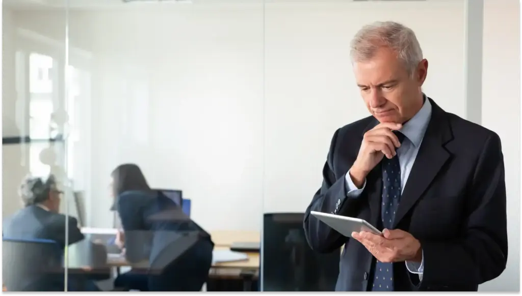 Um chefe de terno está em um escritório segurando um tablet, com uma expressão pensativa, analisando lacuna de autoridade; dois colegas conversam em uma mesa ao fundo.
