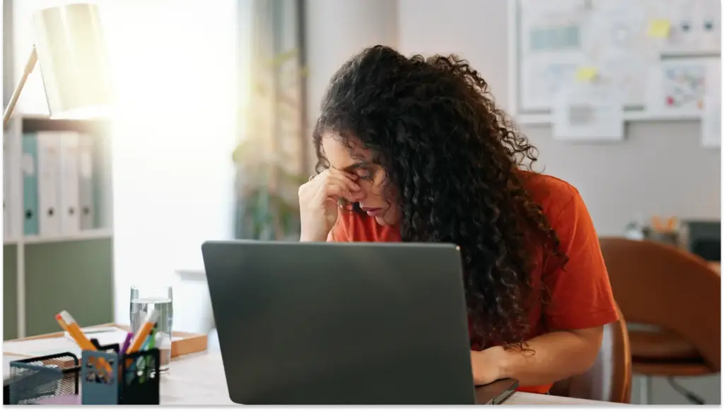 Mulher com cabelo cacheado e expressão de frustração por falta de reconhecimento usando um notebook em um escritório, indicando cansaço ou preocupação.
