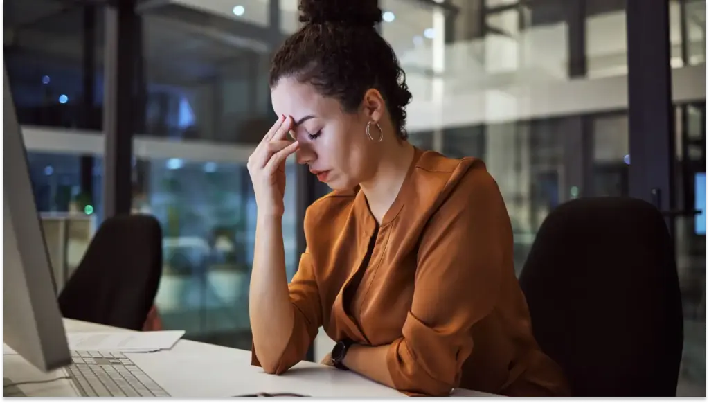 Mulher com expressão de preocupação e frustração, sentada em uma mesa de escritório com computador portátil, lidando com dificuldades da implementação da nova NR-01.