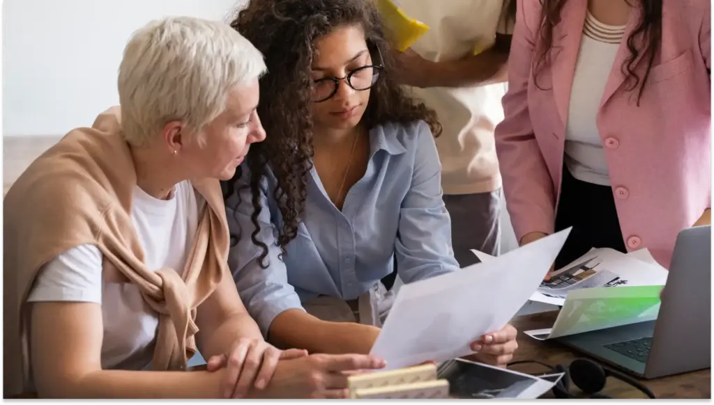 Mulheres de diferentes idades trabalhando juntas em uma reunião, analisando documentos e discutindo estratégias de negócios ou projetos. Mulheres recebem menos salário em comparação aos homens.