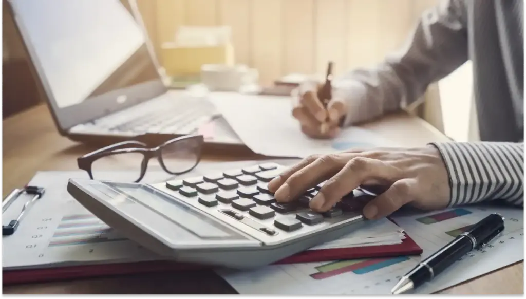 Pessoa utilizando calculadora, calculando Isenção do IR, em uma mesa de escritório com papéis, computador e óculos ao redor, ideal para quem busca dicas de cálculo financeiro ou contabilidade.