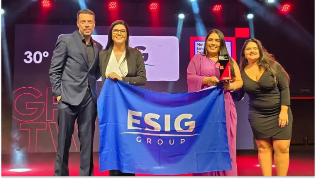 Grupo de pessoas na cerimônia de premiação da ESIG Group, com destaque para uma mulher recebendo um troféu no palco, vestido em rosa, e outras duas mulheres e um homem ao lado, de terno e vestido escuro, em fundo com iluminação artística.