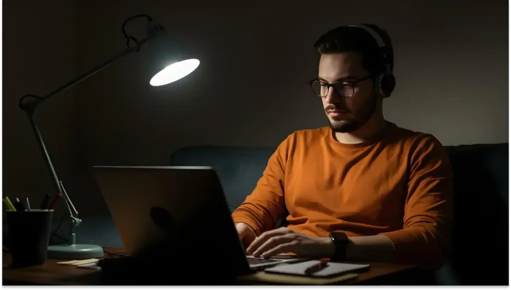 Homem estudando CNPJ ou trabalhando no computador em um ambiente com iluminação suave e foco na tela, usando fones de ouvido e óculos.