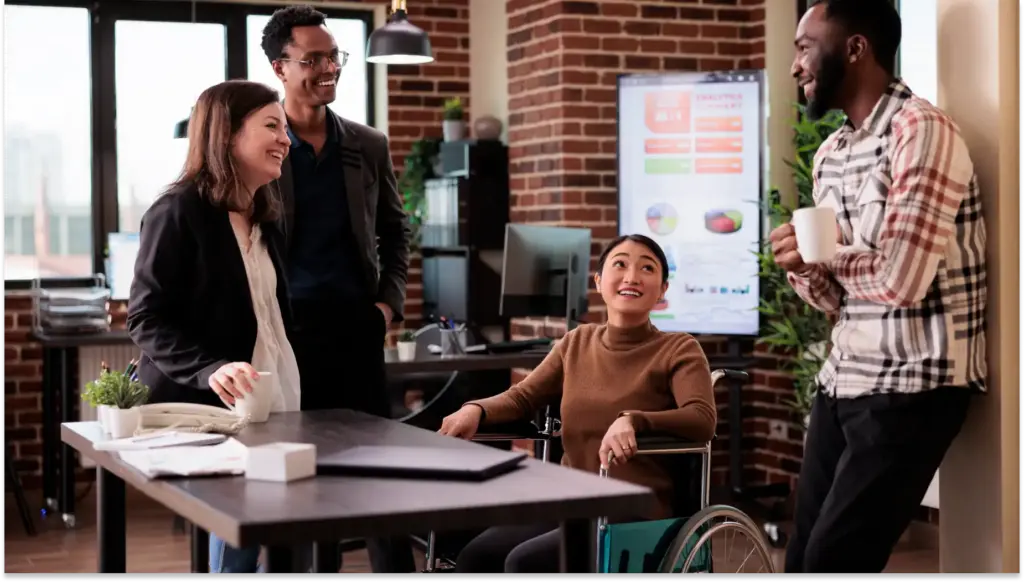 Equipe de trabalho colaborando em ambiente de escritório moderno e inclusivo com uma pessoa em cadeira de rodas, discutindo ideias com colegas, promovendo diversidade e inclusão no local de trabalho.