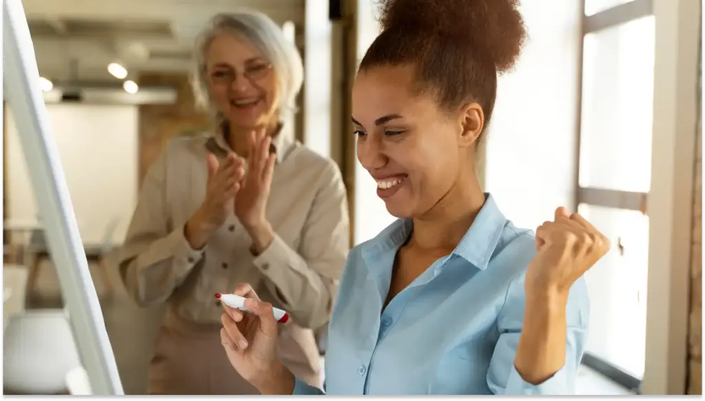 Mulher jovem comemorando benefícios personalizados ao lado de mulher mais velha, ambas sorrindo na frente de um computador em um ambiente de escritório.