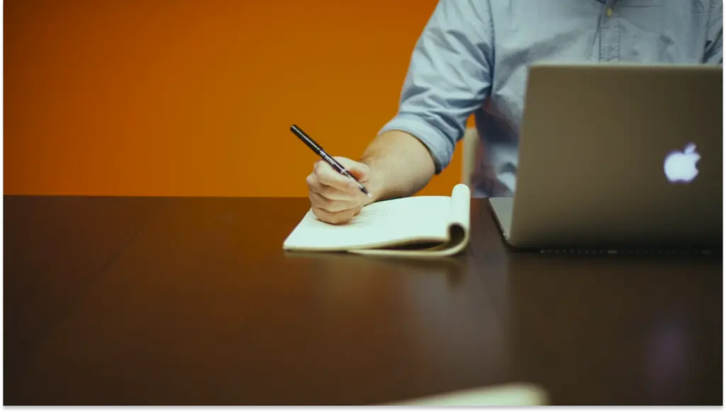 Pessoa escrevendo em caderno ao lado de laptop logado no relatório de transparência salarial 2026, cenário de trabalho ou estudo, no ambiente corporativo com fundo laranja.