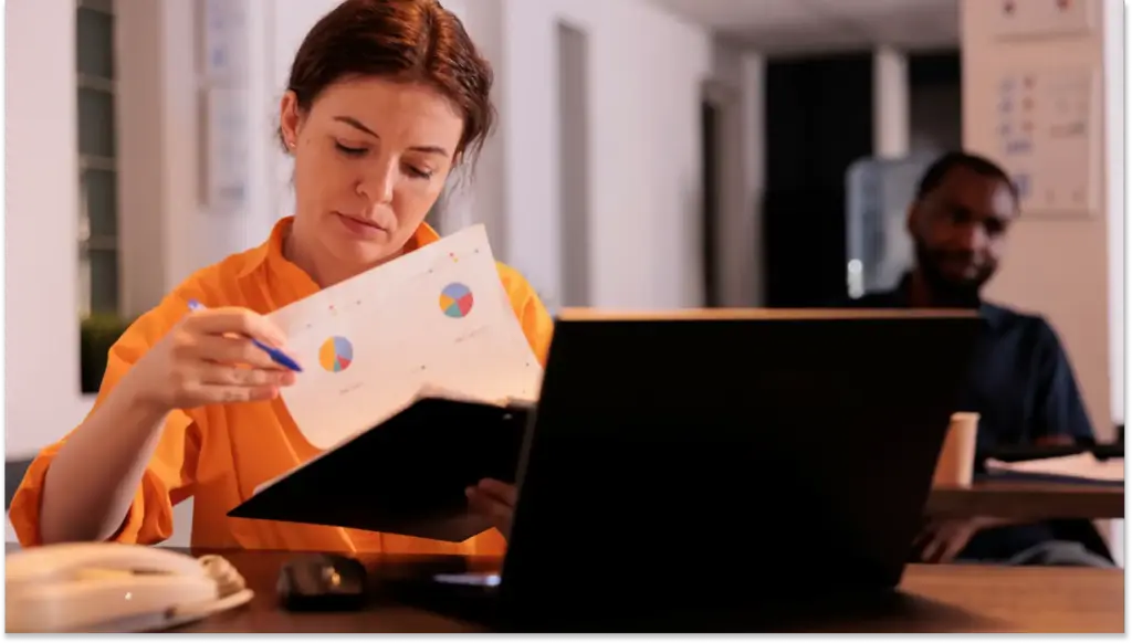 Mulher concentrada analisando gráficos de alta taxa de turnover em uma sala de reunião, com dois colegas ao fundo, numa ambiente de escritório profissional.