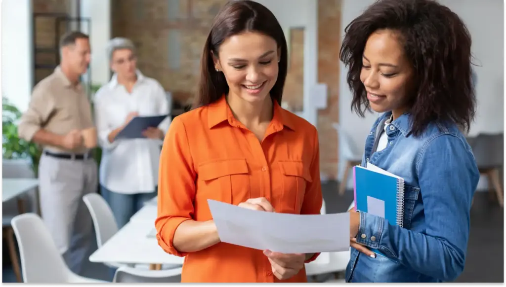 Duas mulheres conversando sobre novos empregos formais e trocando documentos em um ambiente de escritório, enquanto outros colegas discutem ao fundo, simbolizando colaboração e profissionalismo.
