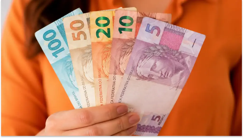 Imagem de mãos segurando diferentes cédulas de dinheiro, representando impacto do reajuste salarial na margem para CLT em 2026.