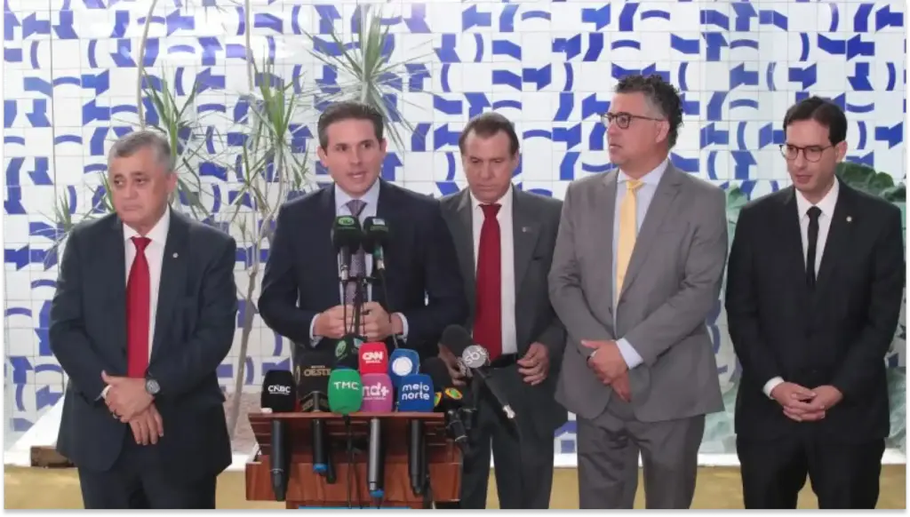 Encontro de autoridades em entrevista coletiva: cinco homens em trajes formais ficam ao lado de mesa com microfones e bandejas, diante de painel com padrão azul e branco, em ambiente externo, disutindo escala 6x1.