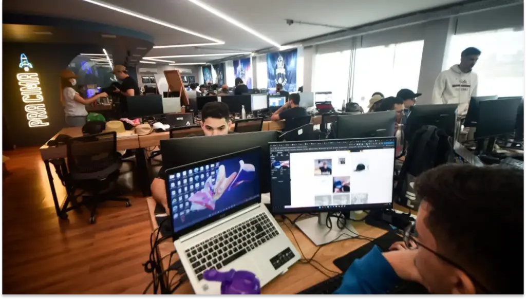 Ambiente de um escritório de tecnologia com várias pessoas trabalhando em computadores, incluindo um espaço de coworking com decoração moderna. QuarkRH