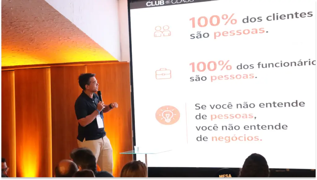 Palestrante em evento corporativo apresentando insights sobre contratação e retenção, com slide “Insights palestra Contratação e Retenção - Carbone Class” e mensagem sobre 100% dos clientes e funcionários serem pessoas. Plateia ao fundo.