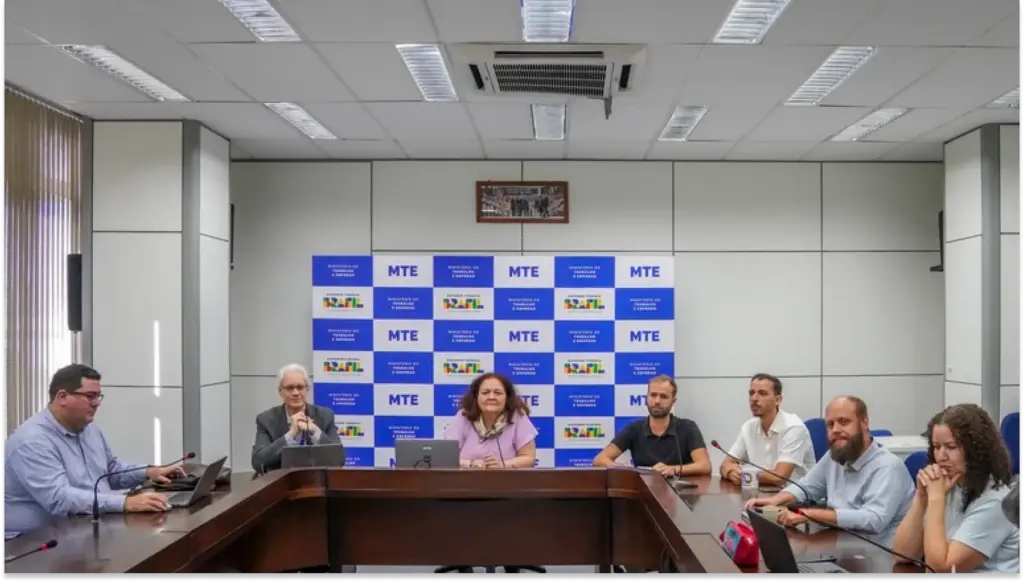 Reunião em sala de comitê do MTE com participantes sentados ao redor de mesa, em evento com painel de fundo azul escrito MTE e monitores, registrada durante encontro institucional.
