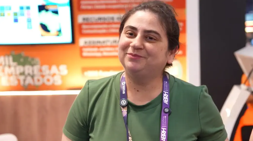 Sevilha: mulher em evento, usando crachá e camiseta verde, sentada em ambiente com banners e tela ao fundo.