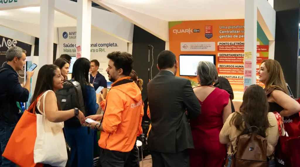 Pessoas em um estande durante o evento corporativo, com funcionários e visitantes conversando e conferindo materiais ao lado de um painel da QuarkRH. QuarkRH participa da 52ª edição do CONARH em SP.