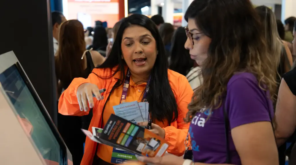 QuarkRH participa da 52ª edição do CONARH em SP: mulher em crachá com camisa laranja apresenta materiais e demonstração em estande durante feira com público ao fundo.