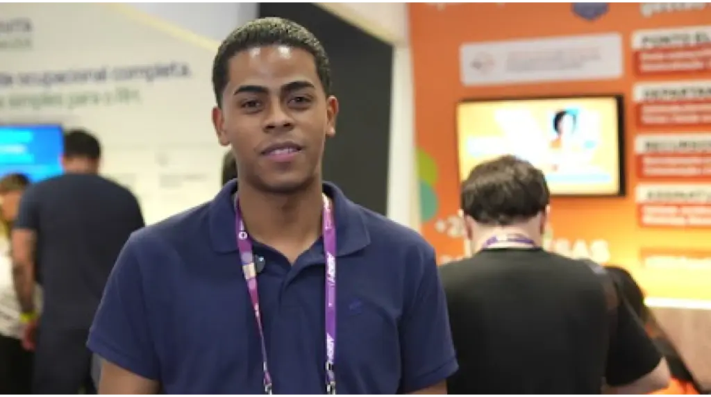 Scientech Ambiental: Participante em evento com crachá e cordão roxo, em frente a um estande com painéis informativos e fundo laranja e amarelo, em ambiente de feira.