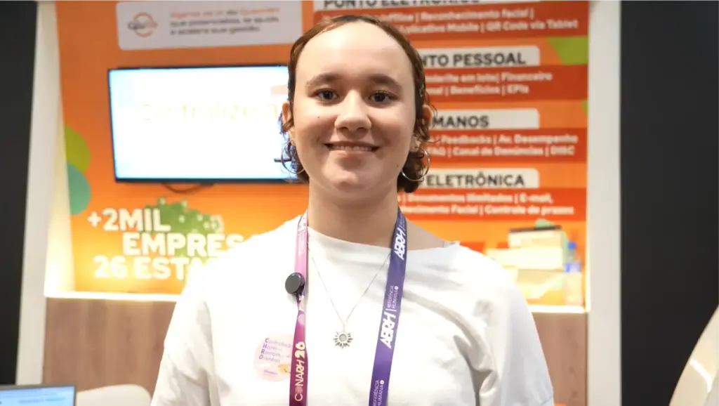 Brigatta: Participante em foto em evento, com crachá e cordão, sorrindo em frente a um painel com informações e números sobre empresas e tecnologia.