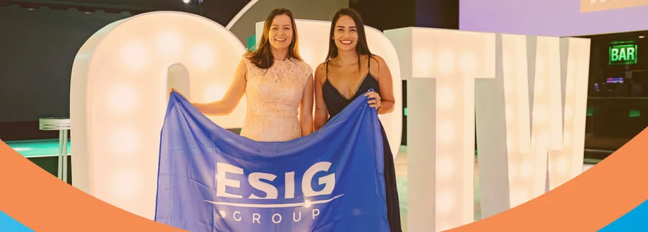 Duas mulheres segurando a bandeira do ESIG Group em um evento festivo, com letras iluminadas ao fundo.