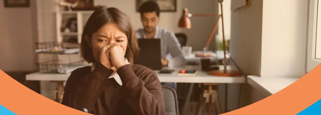 A imagem mostra uma mulher em um ambiente de escritório, expressando preocupação com a saúde, enquanto um homem está ao fundo trabalhando em um computador.