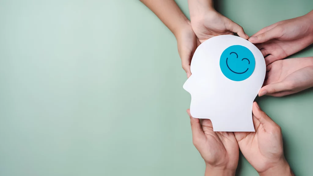 Imagem representativa da saúde mental com mãos segurando uma cabeça de papel com um sorriso, enfatizando sua importância na gestão de pessoas em 2025.