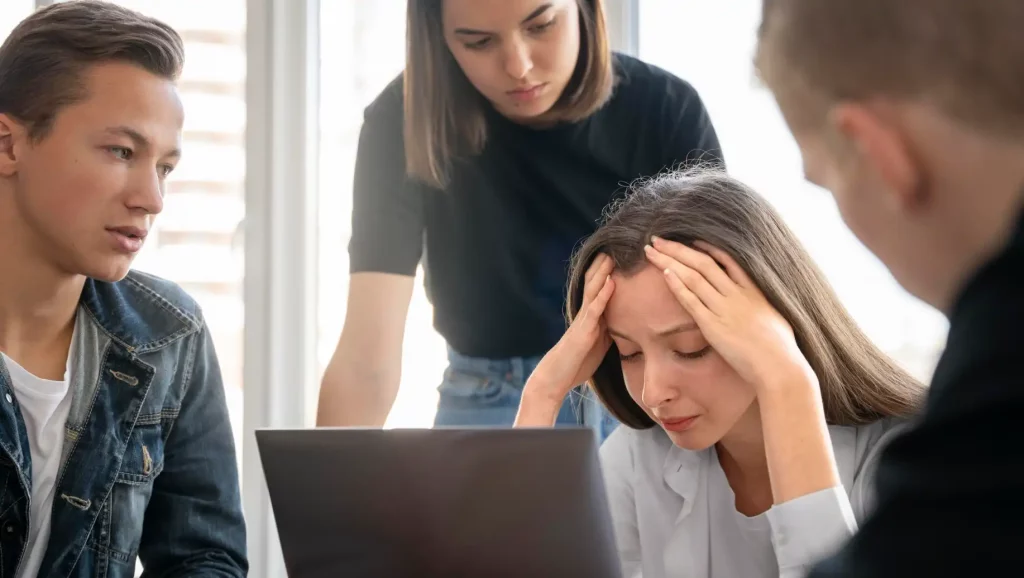 Grupo de jovens planejando demissão voluntária, com destaque para uma jovem demonstrando estresse ao olhar para o laptop. A cena é em um ambiente moderno, favorável ao aprendizado e trabalho em equipe.
