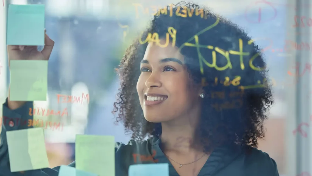 Mulher com cabelos cacheados sorrindo enquanto observa anotações em post-its em uma parede de vidro, transmitindo uma sensação de criatividade e inspiração para tendências de RH.