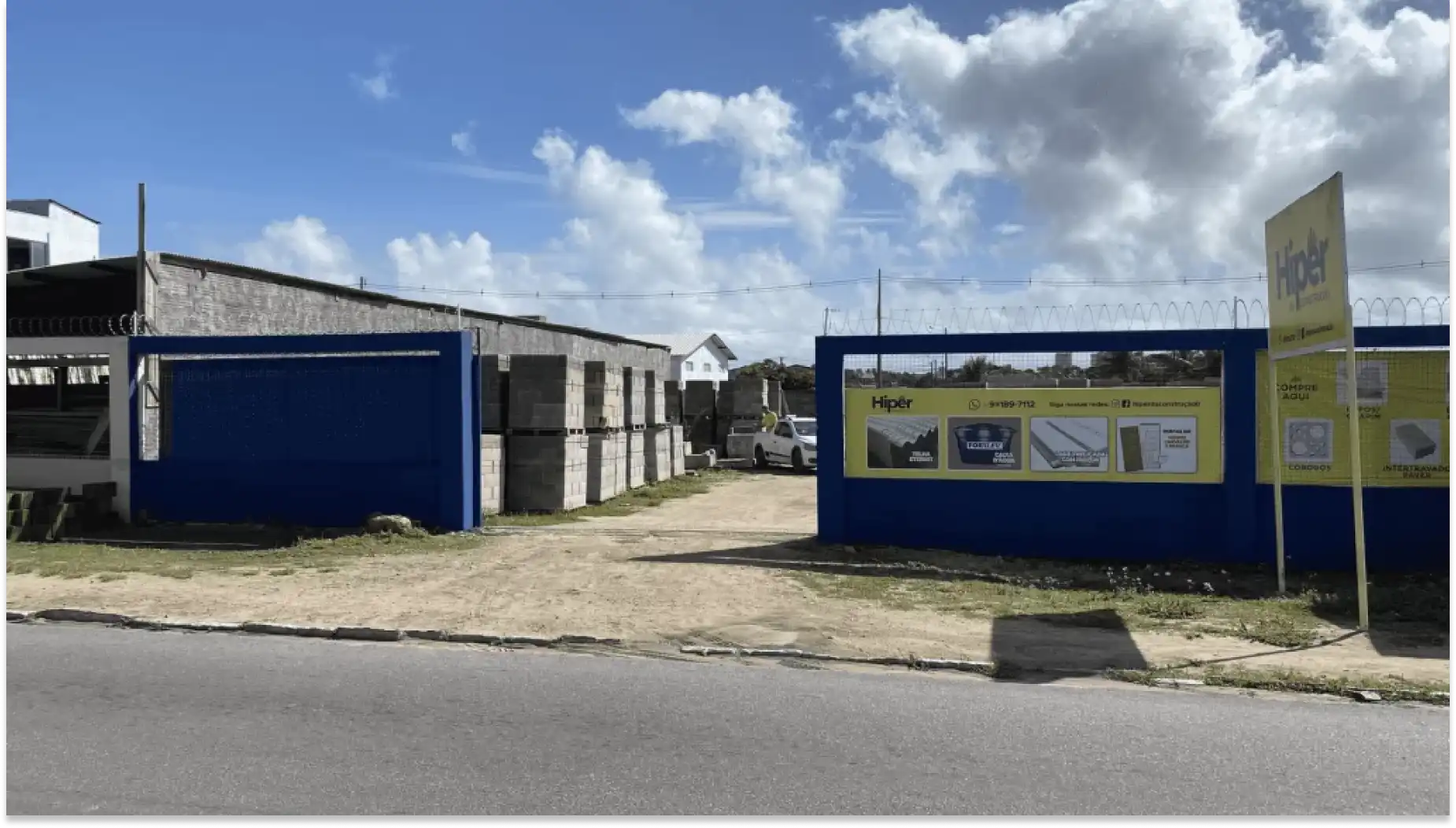 Foto de uma loja de materiais de construção chamada Hiper da construção, situada na rua com estacionamento ao lado e uma área de areia na frente, sob céu com nuvens.