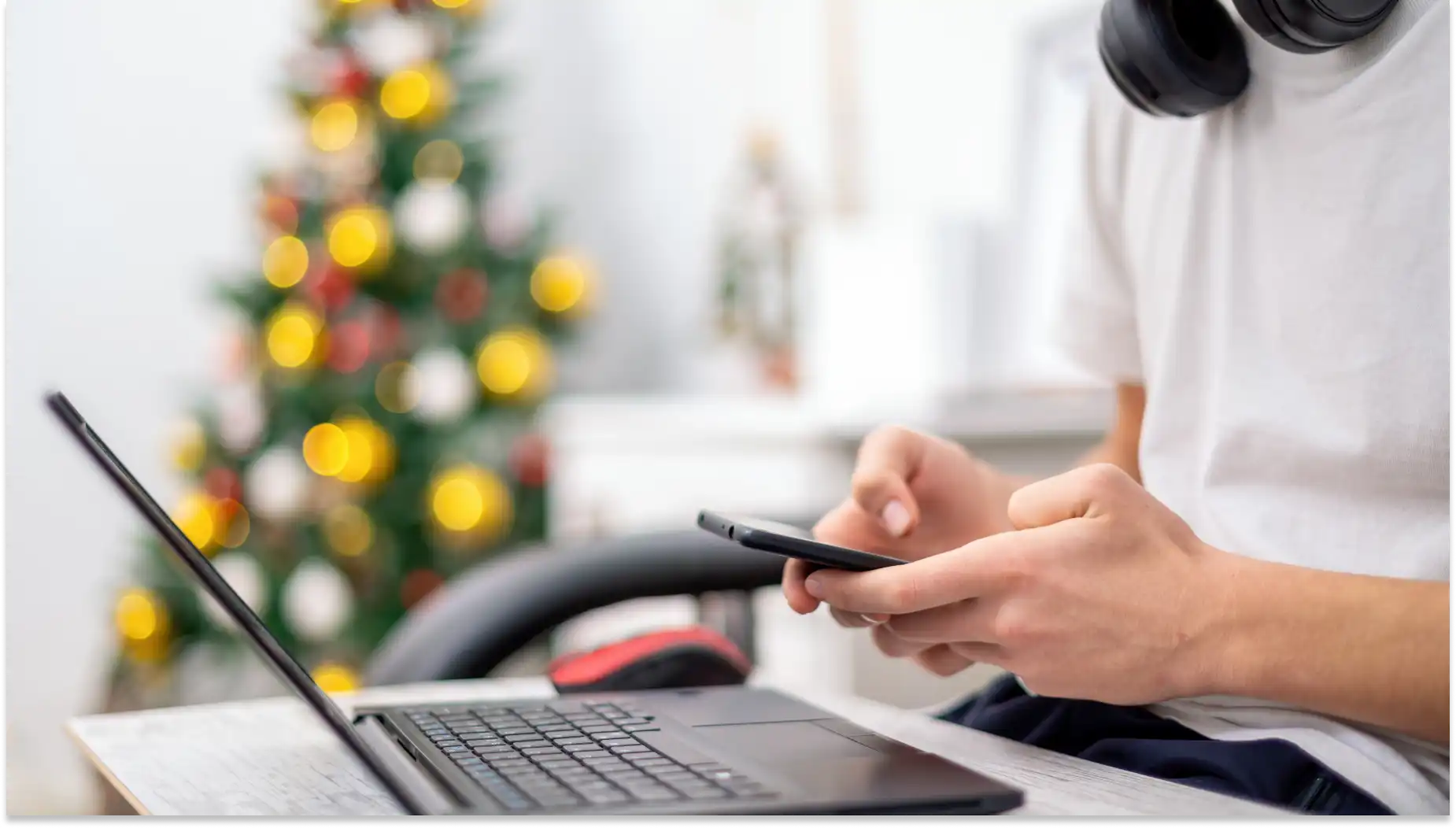 Pessoa usando smartphone em frente ao laptop, com fones de ouvido ao redor do pescoço, ambiente com árvore de Natal decorada ao fundo, preparando para o Vale-Natal.