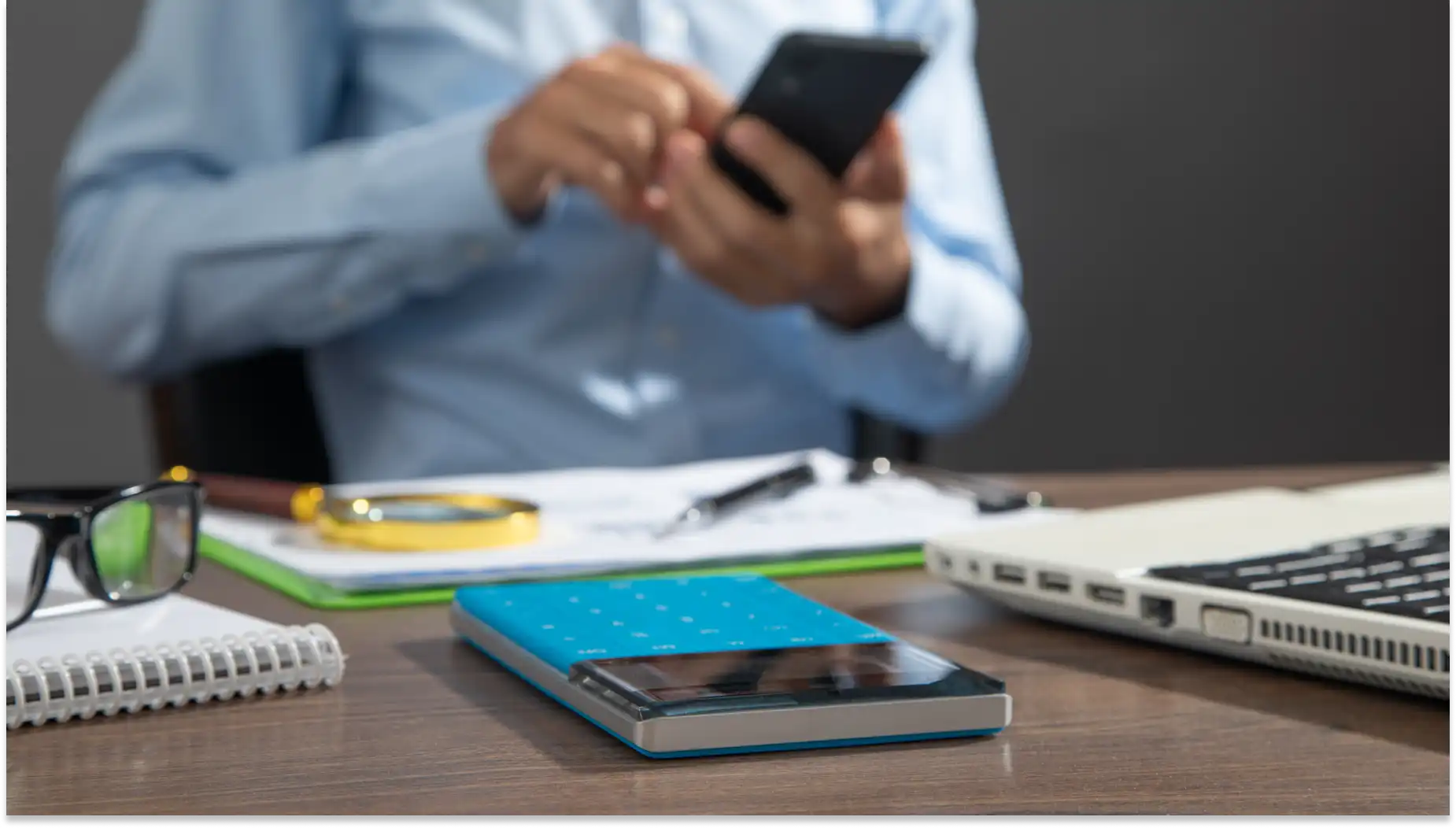 Pessoa usando smartphone em ambiente de escritório com objetos de trabalho na mesa, incluindo notebook, livros, óculos e caderno, em uma atmosfera de produtividade, calculando um empréstimos consignados.