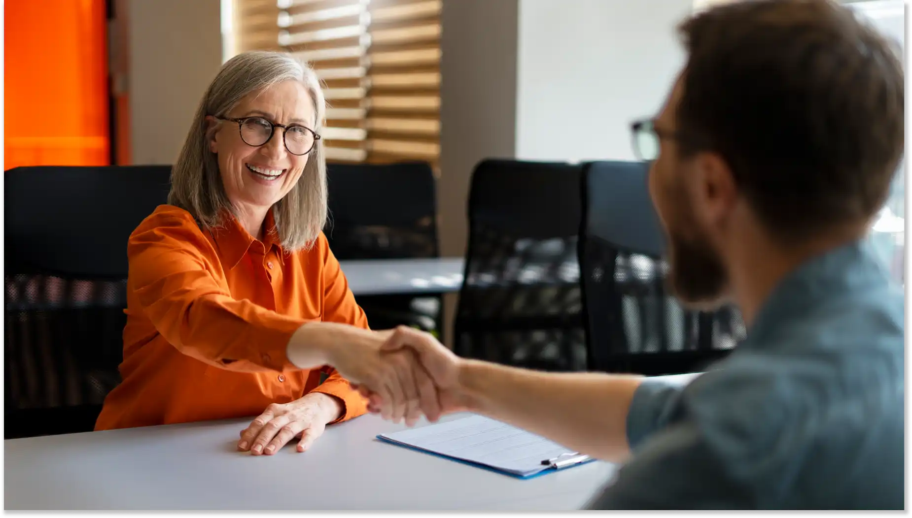 Mulher idosa sorridente dando um aperto de mão a um homem durante uma entrevista de emprego, ambos sentados em uma mesa de escritório com ambiente profissional ao fundo. Novo Caged no Brasil, gerando novos empregos.