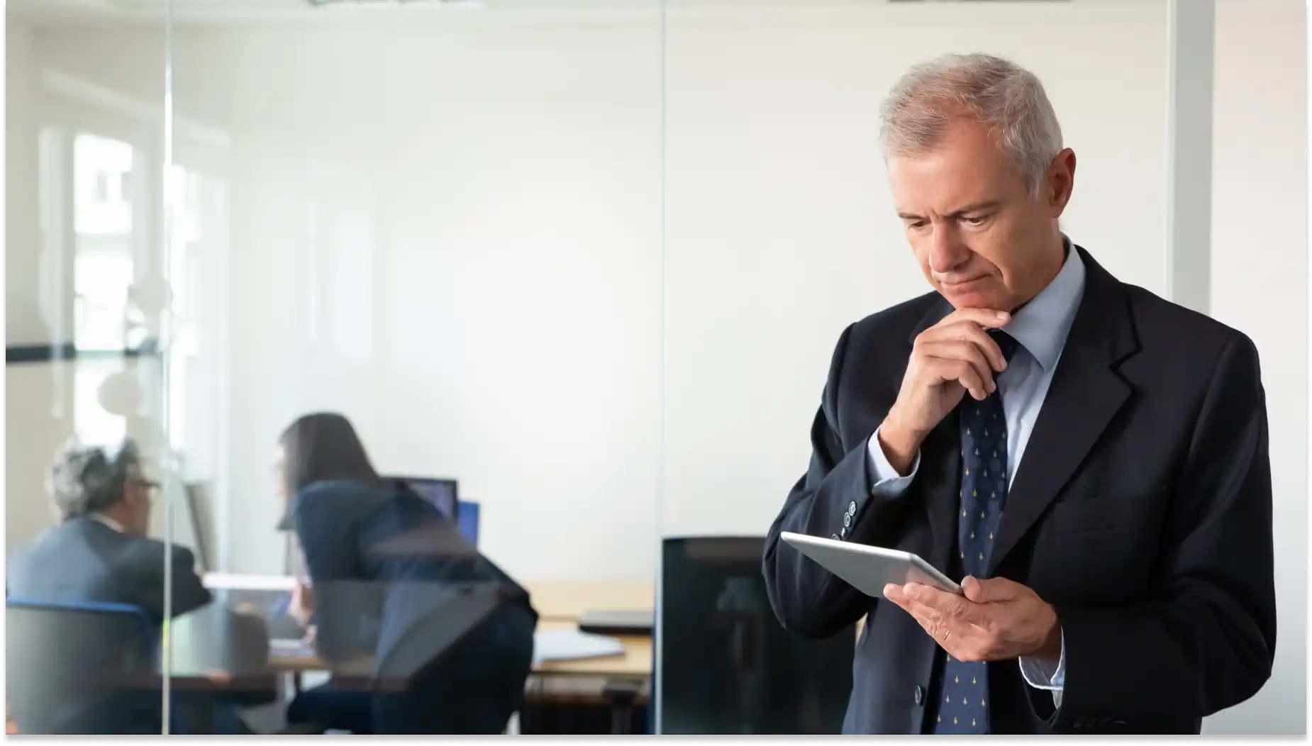 Um chefe de terno está em um escritório segurando um tablet, com uma expressão pensativa, analisando lacuna de autoridade; dois colegas conversam em uma mesa ao fundo.