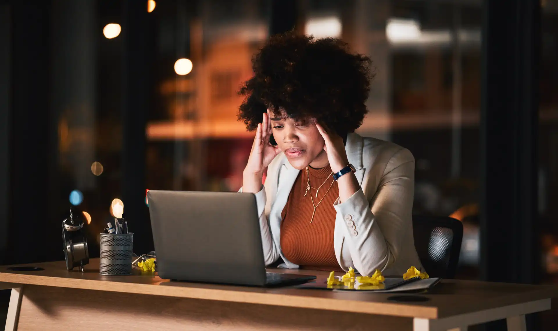Mulher jovem com cabelo cacheado, usando blazer bege e roupa marrom, segurando a cabeça e parecendo ter pouca segurança psicológica enquanto trabalha no computador em um escritório à noite.