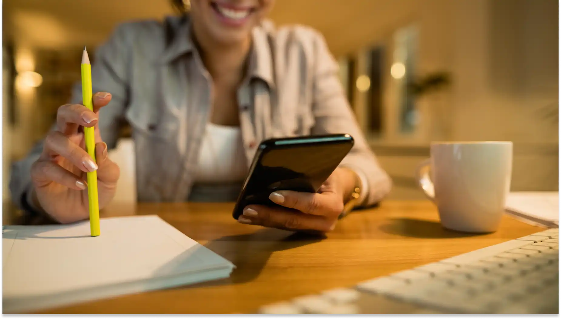 Mulher sorridente usando smartphone em ambiente de trabalho com caneta, caderno e caneca na mesa, calculando a nova faixa de isenção do IR, promovendo produtividade e comunicação eficaz.
