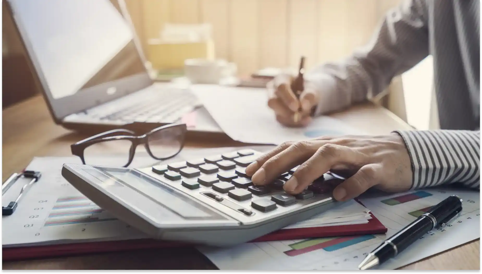 Pessoa utilizando calculadora, calculando Isenção do IR, em uma mesa de escritório com papéis, computador e óculos ao redor, ideal para quem busca dicas de cálculo financeiro ou contabilidade.