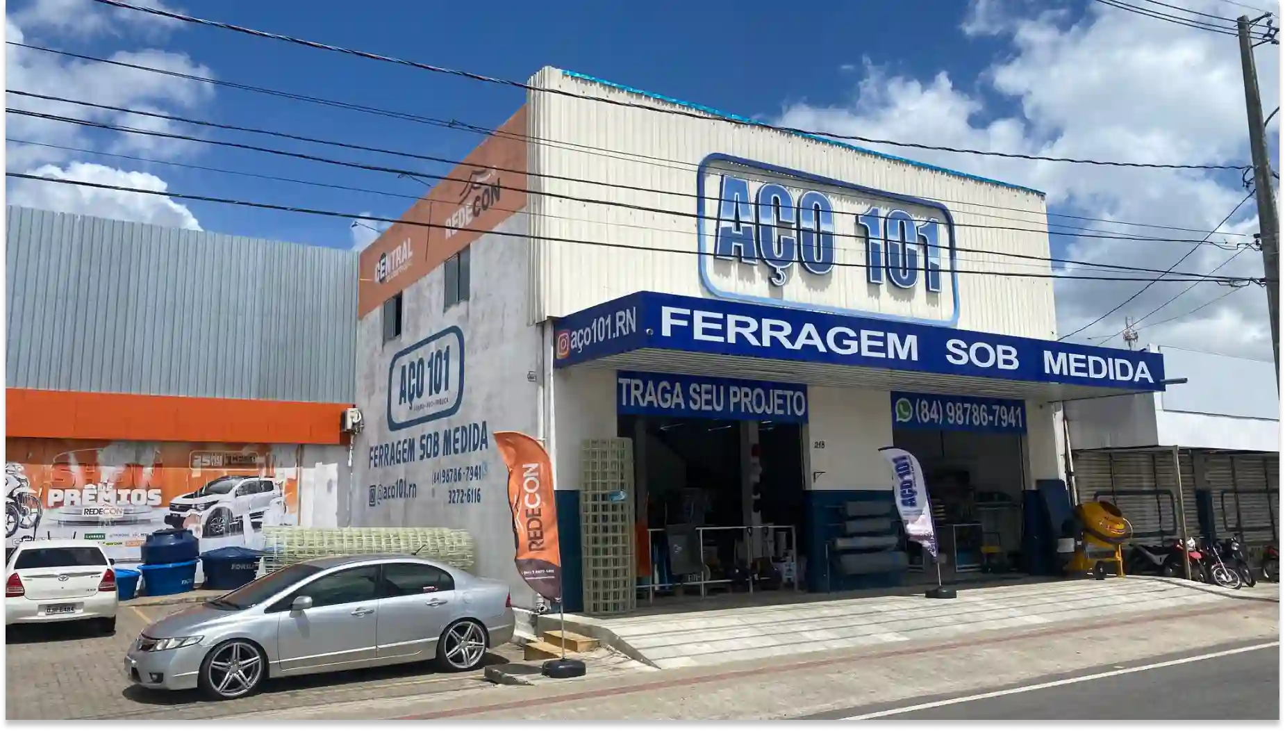 Fachada da loja Aço 101 especializada em ferragem sob medida, com veículos estacionados na frente e um aviso de serviço personalizado.