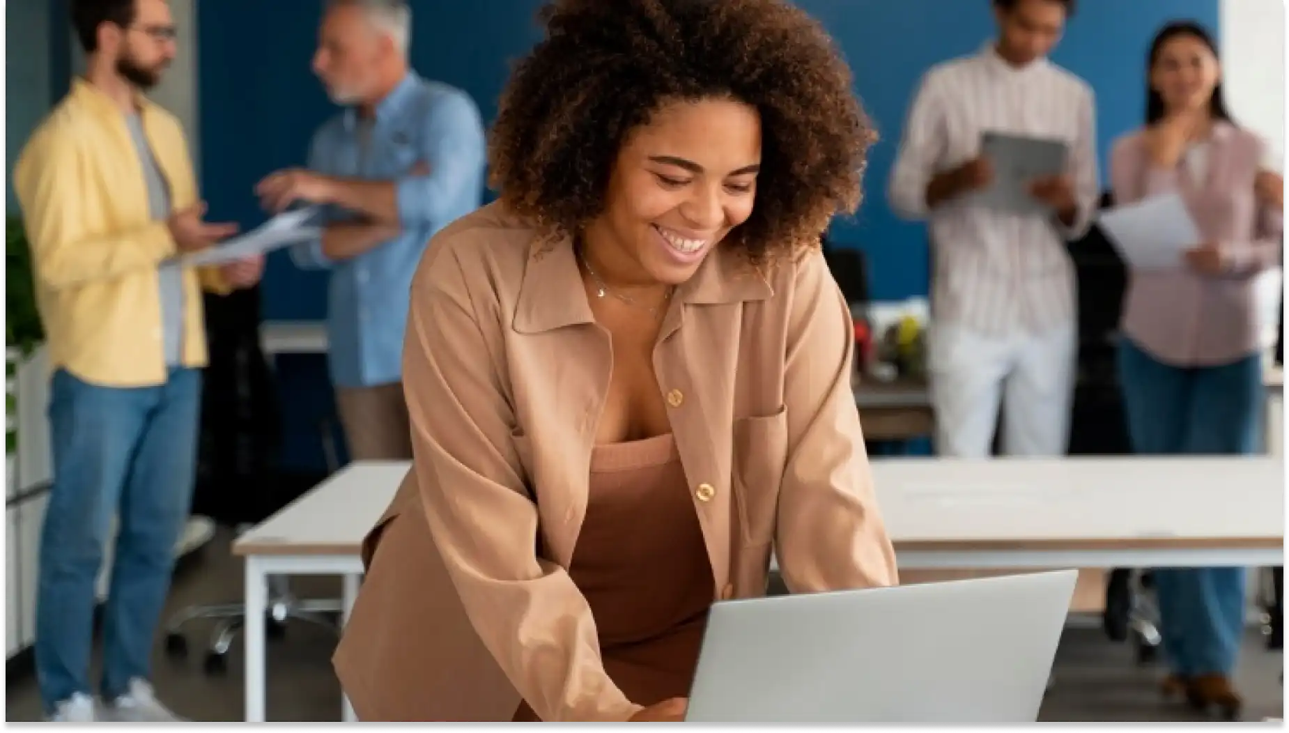 Mulher sorrindo por conta do Fim da Escala 6X1, usando um computador portátil em ambiente de escritório com colegas ao fundo